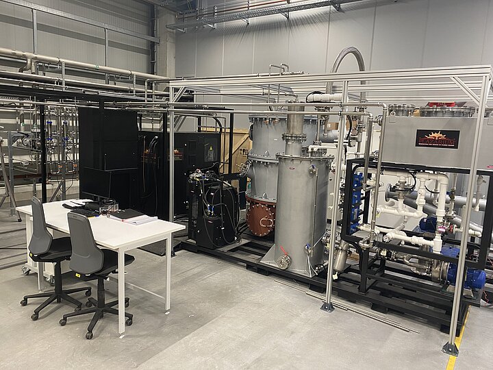 Zdjęcie przedstawiające laboratorium do testów plazmowych. W centralnej części hali widoczne są stalowe komory do badań plazmowych, generator plazmy oraz układy pomp. Z lewej strony umieszczony jest stół z dwoma krzesłami. Photo showing the plasma testing laboratory. The central part of the hall shows the steel plasma test chambers, the plasma generator and the pump systems. A table with two chairs is placed on the left.