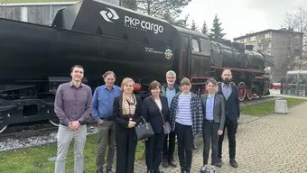 Zdjęcie grupy 8 osób na tle lokomotywy wykonane przy budynku B5 na terenie AGH. Photo of a group of 8 people in front of a locomotive taken by the B5 building on the AGH grounds.
